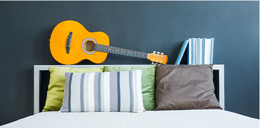 Bedroom with navy wall and bed with colorful cushions and a box headboard that functions as a shelf with books and an acoustic guitar